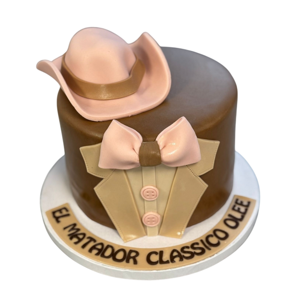 "Runde Torte mit braunem Fondant, dekoriert mit einem Matador-Hut, einer Fliege und einem eleganten Anzug. Schriftzug 'El Matador Classico Olee' auf der Basis. Perfekt für Themenpartys oder elegante Feiern."