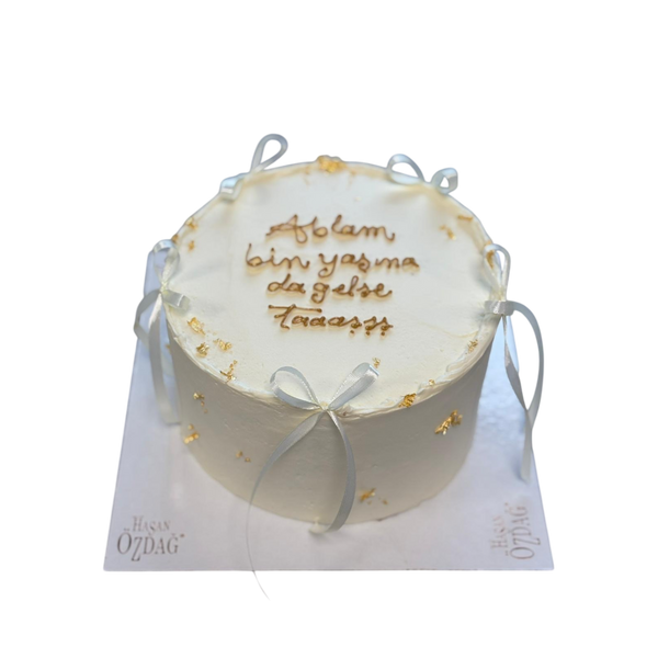 "Runde Torte in Weiß, dekoriert mit goldenen essbaren Akzenten, zarten Schleifen und der personalisierten Botschaft 'Ablam bin yaşa da gelse taş'. Perfekt für Geburtstage oder besondere Anlässe."