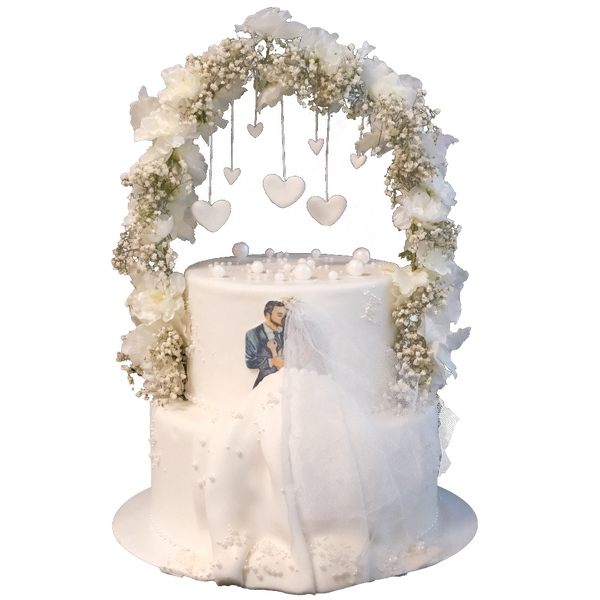 Weiße zweistöckige Hochzeitstorte mit Blumen-Bogen, an dem Herzen hängen.  Vorne ist eine Brautpaar-Illustration mit 3D-Kleid aus Fondant. Die Torte ist mit Perlen dekoriert.