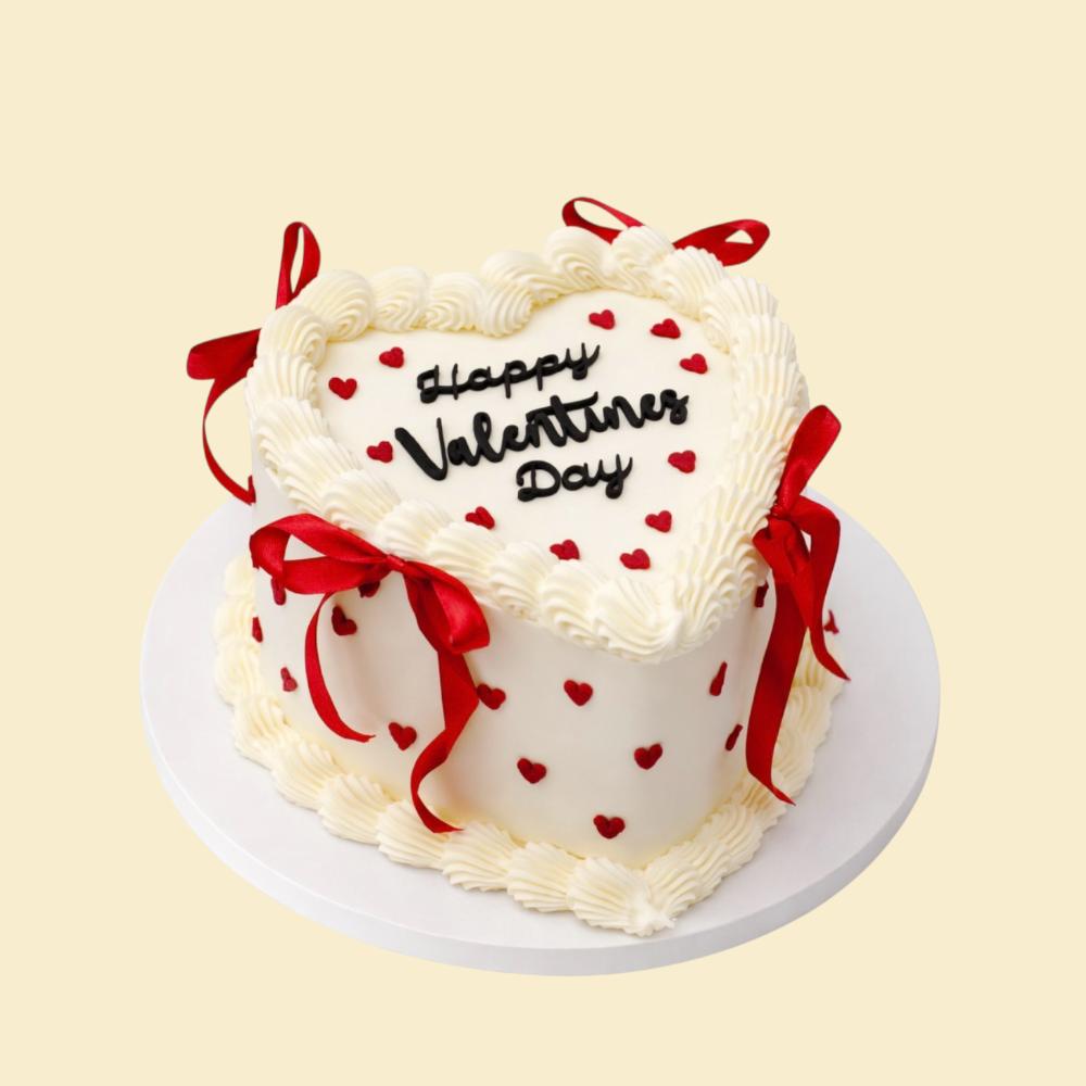 Herzförmige Valentinstagstorte in Weiß mit roter Schleifendekoration, kleinen roten Herzen und der Aufschrift „Happy Valentines Day“ auf der Oberseite.