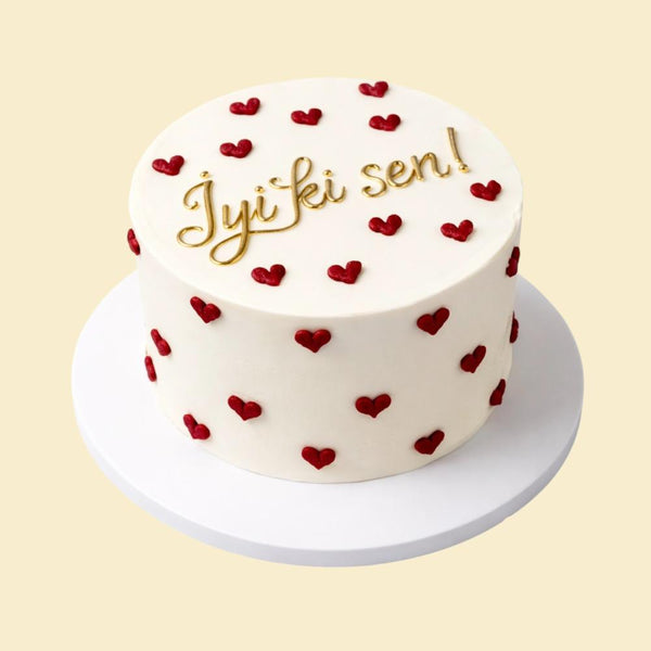 Runde weiße Torte mit kleinen roten Herzdekorationen rundum und der goldenen Aufschrift „İyi ki sen!“ auf der Oberseite, präsentiert auf einer weißen Tortenplatte.