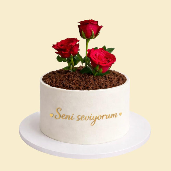 Weiße, runde Torte mit realistischer Erdoptik auf der Oberfläche und drei roten Rosen, verziert mit der goldenen Aufschrift „Seni seviyorum“, präsentiert auf einer weißen Tortenplatte.