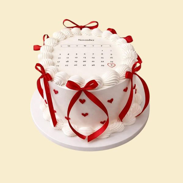 Weiße Torte mit Kalenderdesign, dekoriert mit feinen Buttercreme-Wellen, roten Schleifen und kleinen Herzdetails; ein Datum ist rot markiert und hervorgehoben, arrangiert auf einer weißen Tortenplatte.