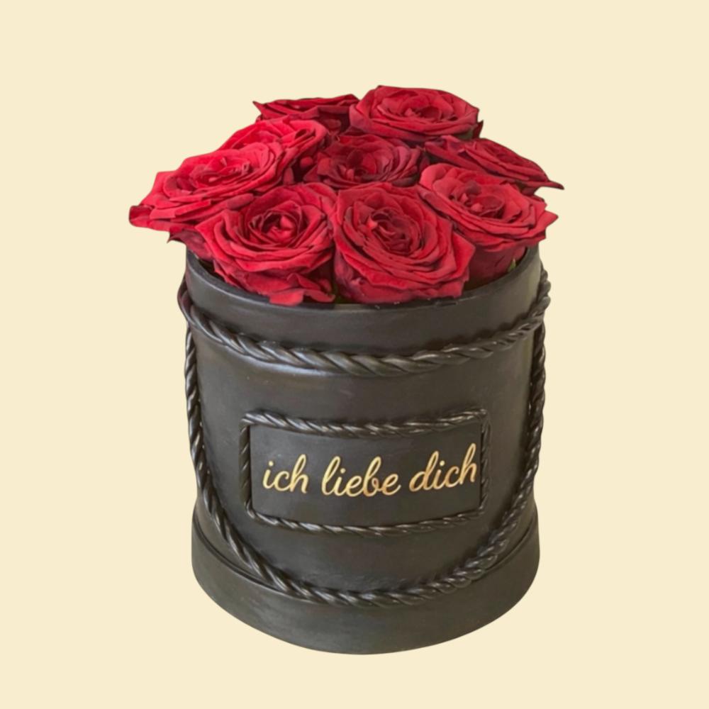 Schwarze zylindrische Torte im luxuriösen Design, gefüllt mit roten Rosen und verziert mit einer goldenen Aufschrift „ich liebe dich“ auf der Vorderseite; präsentiert vor hellem Hintergrund.