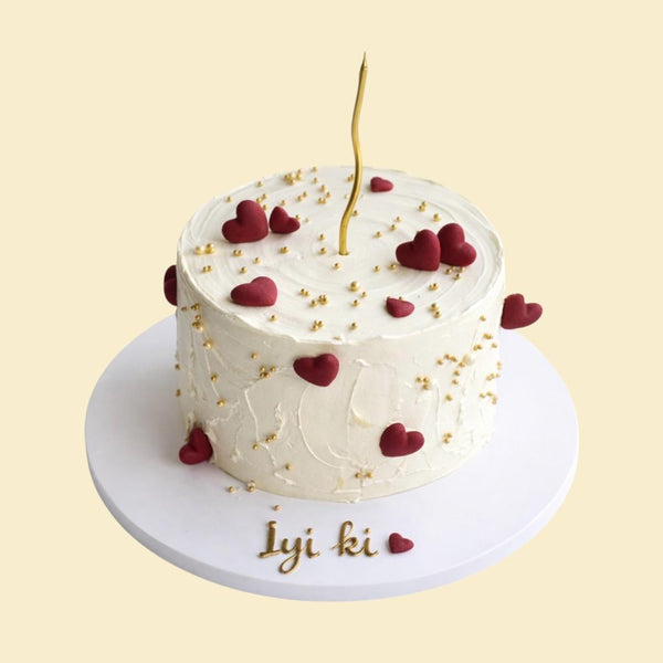 Weiße, runde Torte mit glatter Creme Oberfläche, dekoriert mit roten Herzmotiven und goldenen Zuckerperlen. Oben steht eine einzelne goldene Kerze. Auf dem Tortenbrett ist die Aufschrift „İyi ki“ mit einem kleinen roten Herz zu sehen.