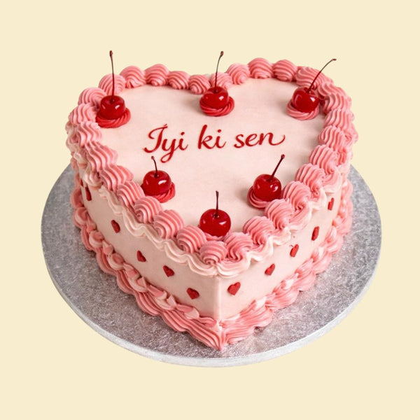 Herzförmige Torte in zarten Rosatönen mit aufgespritzter Creme, dekoriert mit roten Kirschen und kleinen Herzdetails, beschriftet mit „İyi ki sen“, auf silberner Tortenplatte vor hellem Hintergrund.