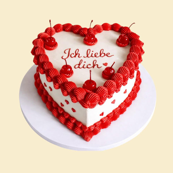 Herzförmige Torte mit weißer Oberfläche, roter Spritzrand Dekoration und Kirschen, mit der Aufschrift „Ich liebe dich“ auf einer weißen Tortenplatte.