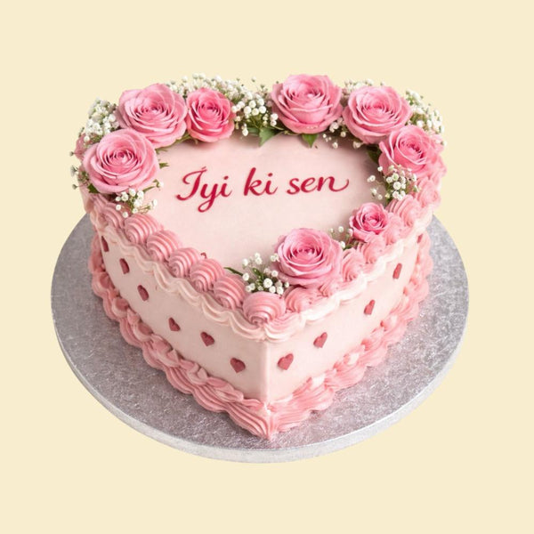 Herzförmige rosafarbene Torte mit feiner Spritzdekoration, kleinen Herzdetails und einem Kranz aus rosa Rosen und Schleierkraut, mit der Aufschrift „İyi ki sen“ auf einer silbernen Tortenplatte.