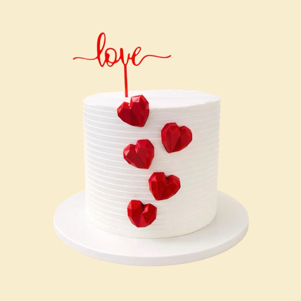 Weiße, moderne Valentinstorte mit gerillter Oberfläche, roten geometrischen Herzen an der Seite und rotem „love“-Cake-Topper auf einer runden weißen Tortenplatte.