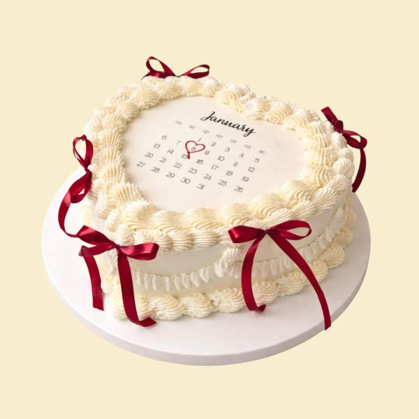 Herzförmige Kalendertorte in Cremeweiß mit Januar-Kalender, markiertem Datum, feinen Buttercreme-Verzierungen und roten Schleifen auf weißer Tortenplatte