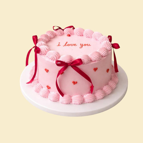 Rosa Valentinstorte mit Cremebordüre, roten Schleifen, kleinen Herzdetails und der Aufschrift „I love you“ auf einer weißen Tortenplatte.