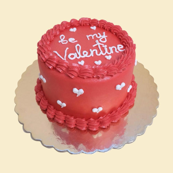Rote Valentinstorte mit Cremebordüre, kleinen weißen Herzen und der Aufschrift „Be my Valentine“ auf silberner Tortenplatte.