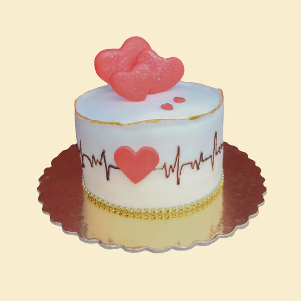 Weiße, runde Torte mit Herz-EKG-Motiv, rosafarbenen Herzdekoren auf der Oberseite und goldenen Akzenten auf goldener Tortenplatte.


