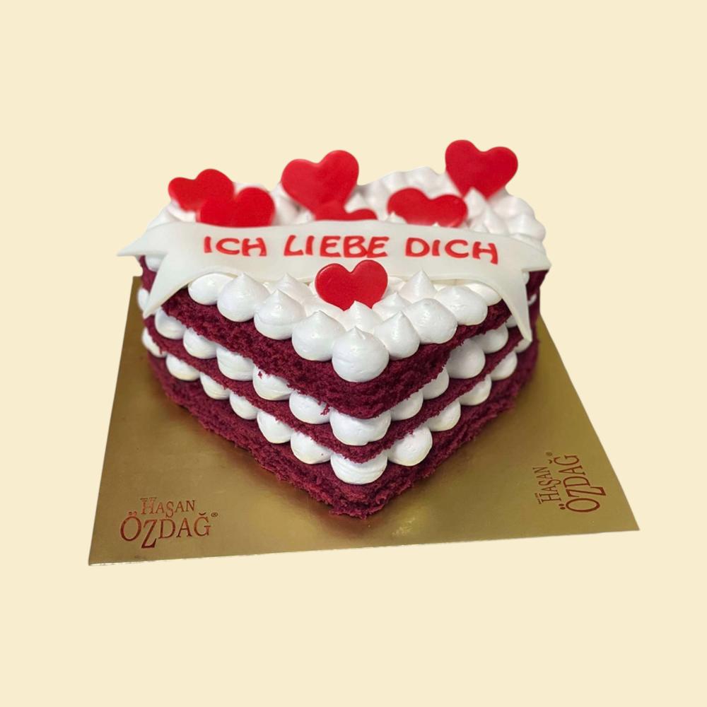 Herzförmige Red-Velvet-Torte mit mehreren Schichten weißer Creme, roten Herzdekorationen und Schriftzug „Ich liebe dich“ auf goldener Tortenplatte.