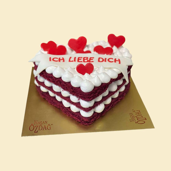Herzförmige Red-Velvet-Torte mit mehreren Schichten weißer Creme, roten Herzdekorationen und Schriftzug „Ich liebe dich“ auf goldener Tortenplatte.