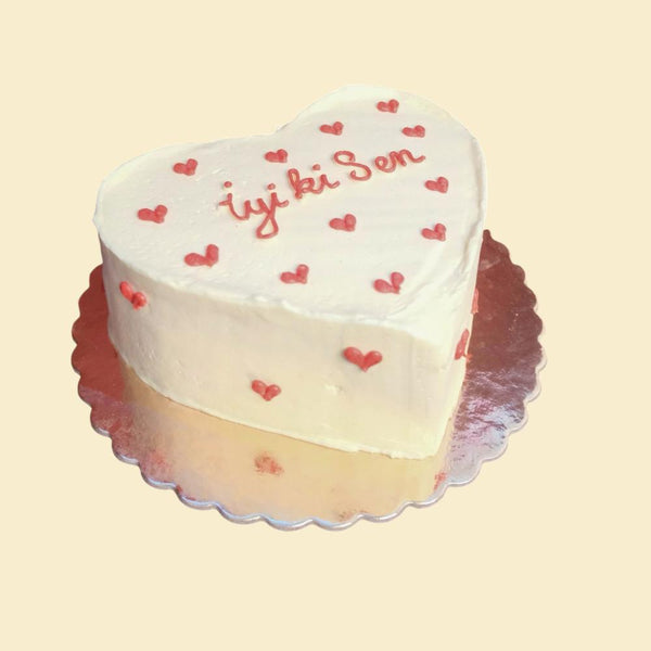 Herzförmige Torte mit glatter weißer Creme, kleinen roten Herzmotiven und handgeschriebenem Schriftzug „İyi ki sen“ auf silberner Tortenplatte.