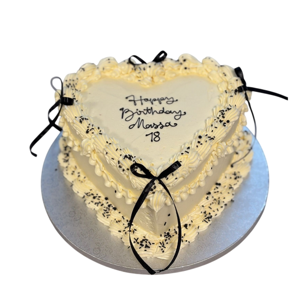 "Elegante Herztorte in Weiß, dekoriert mit feiner Buttercreme, schwarzen Schleifen und einem personalisierten Schriftzug 'Happy Birthday Massa 18'. Perfekt für Geburtstage, Jubiläen und besondere Anlässe."